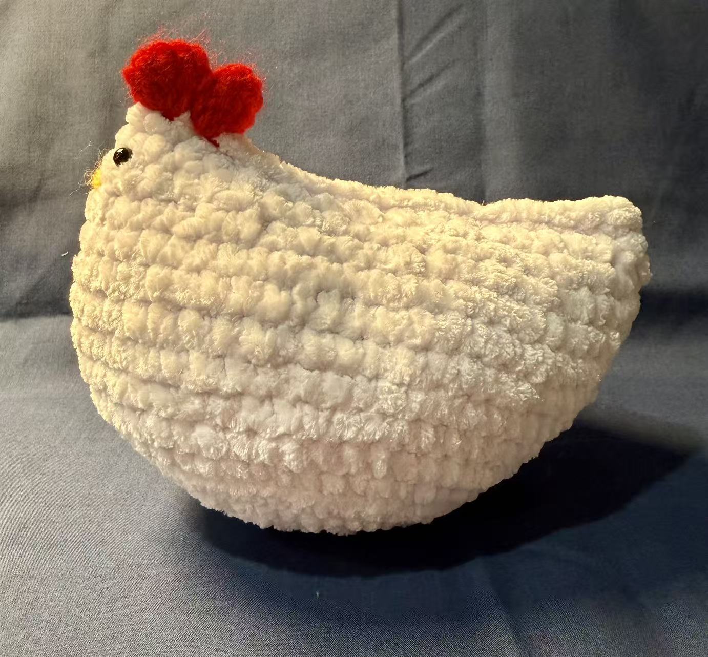 Side view of the white crochet chicken with two black eyes, a yellow stitched beak, and a red comb.