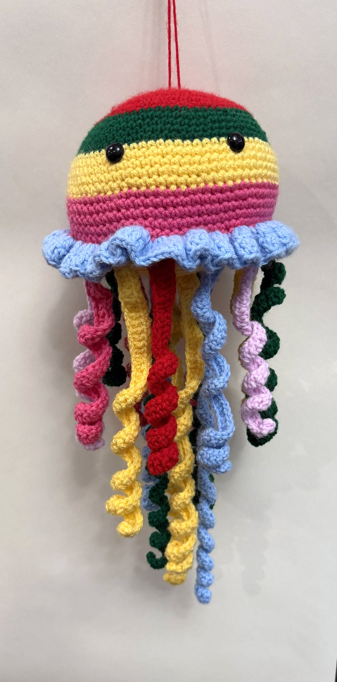 Handmade yarn jellyfish with a striped red, green, yellow, and pink head, black bead eyes, a blue ruffle, and colorful curled strands.
